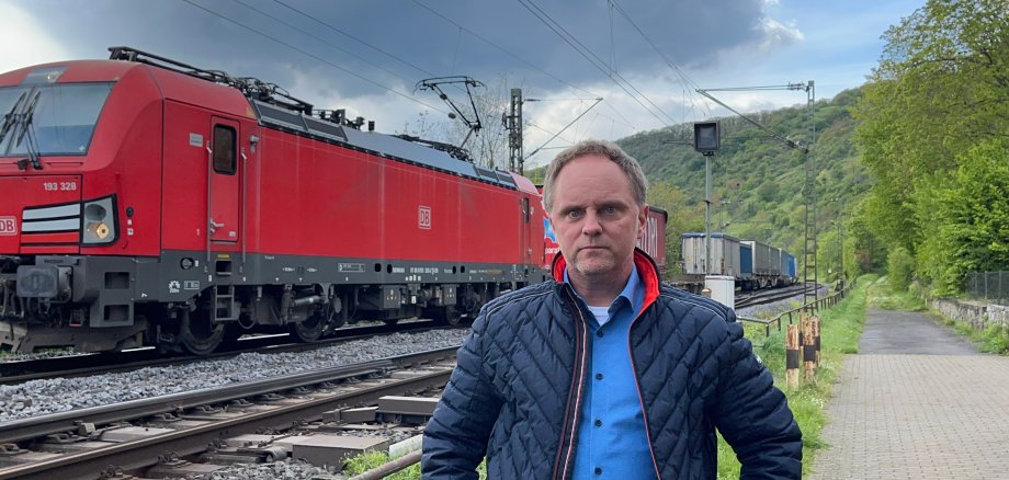 Mike Weiland steht an der Bahnstrecke im Mittelrheintal während ein Güterzug im Hintergrund vorbeifährt