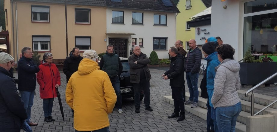 Bürgermeister Mike Weiland und circa 20 weitere Personen stehen bei bewölktem Wetter im Kreis auf dem Dorfplatz in Filsen. 