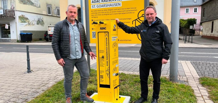 Verwaltungsmitarbeiter Armin Schaust und Bürgermeister Mike Weiland stehen neben der neben der Radreparatursäule.  Dahinter befindet sich ein ebenfalls gelbes Schild, dass auf die Zusammenarbeit mit dem ADAC-Mittelrhein hinweist. Im Hintergrund sind die Straße und weitere Gebäude zu sehen.