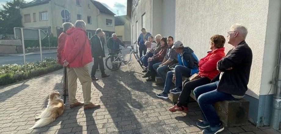 Etwa 15 Menschen sitzen nebeneinander auf einer Bank. Ein paar weitere stehen neben Bürgermeister Mike Weiland, der gerade etwas am vortragen ist.