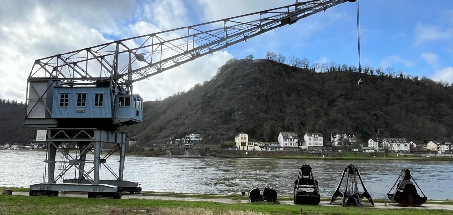 Es ist der alte Schiffs-Kran in St. Goarshausen am Rhein zu sehen. Außerdem sieht man vier Kranschaufeln, die auf dem Boden liegen. Das Wetter ist bewölkt.