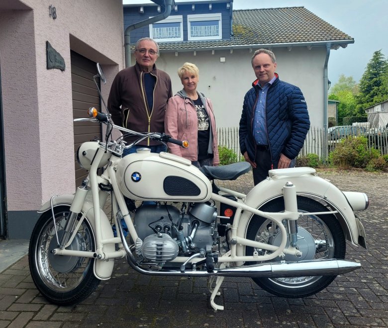 Museumsinhaber Reinhard Gaede, Gisela Adam und Bürgermeister Mike Weiland stehen hinter einem weißen BMW-Oldtimermotorrad. Im Hintergrund ist ein Haus mit Garten zu sehen.