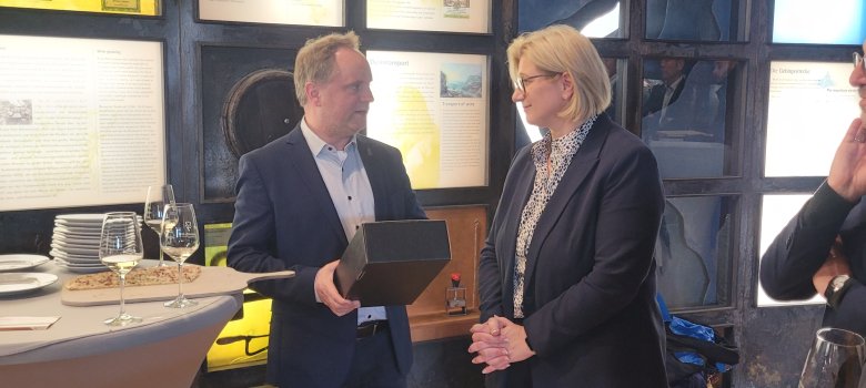 Bürgermeiser Mike Weiland überreicht eine schwarze Box, in der sich eine Flasche Wein befindet, an Saarlands Ministerpräsidentin Anke Rehlinger.