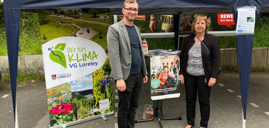 VG-Klimaschutzmanager Hussam Shahoud Alnajm und eine interessierte Bürgerin vor dem Info-Stand, ein blauer Aufstellpavillion, auf dem Parkplatz des REWE-Marktes in St. Goarhausen.