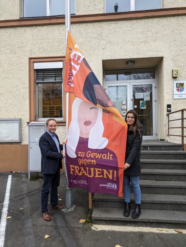 Bürgermeister Mike Weiland und Gleichstellungsbeauftragte Jasmin Fluck hissen gemeinsam die Flagge zum internationalen Tag zur Beseitigung von Gewalt gegen Frauen vor dem Verwaltungsgebäude in St. Goarshausen.